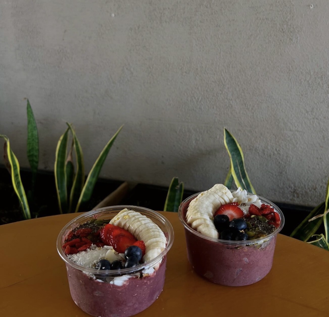 Two açaí bowls on a table with fresh fruit toppings and plants in the background