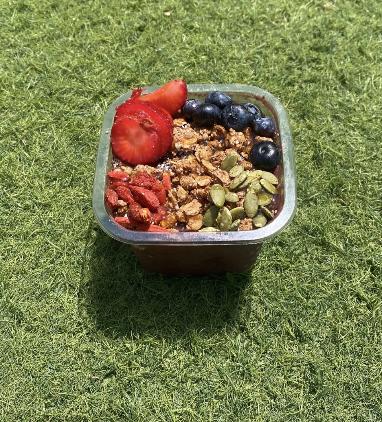 Açaí bowl on grass topped with granola, strawberries, blueberries, and pumpkin seeds